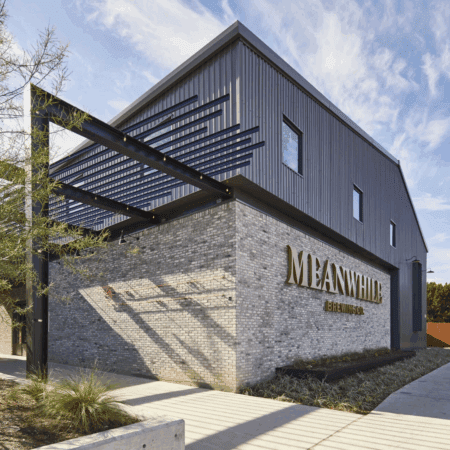 Modern gray brick and metal building with "Meanwhile Brewing Co." sign on the corner, black metal beams overhead, landscaped greenery, and a sidewalk under a partly cloudy sky—a standout among local Austin breweries. Austin Marathon Half Marathon & 5K