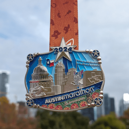 A 2024 Austin Marathon medal featuring a detailed cityscape of Austin landmarks, including the Texas State Capitol, with colorful enamel accents and a star at the top. The belt buckle medal hangs from an orange ribbon adorned with silhouettes of runners against a cloudy sky backdrop. Austin Marathon Half Marathon & 5K A 2024 Austin Marathon medal featuring a detailed cityscape of Austin landmarks, including the Texas State Capitol, with colorful enamel accents and a star at the top. The belt buckle medal hangs from an orange ribbon adorned with silhouettes of runners against a cloudy sky backdrop. Austin Marathon Half Marathon & 5K