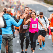 Runners in the marathon push through and exchange high-fives with spectators along the course. A woman in a pink top smiles while engaging with the crowd, embodying the spirit of conquering hard miles. Other runners follow behind, their colorful athletic wear visible against the blurred background that enhances the sense of movement. Austin Marathon Half Marathon & 5K