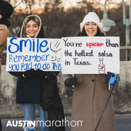 Two people stand outdoors, bundled up in warm clothing, holding humorous signs. One sign says, "Smile. Remember you paid to do this," while the other reads, "You're spicier than the hottest salsa in Texas." Both are smiling. Text reads "Austin Marathon" at the bottom—a celebration of marathon running myths and camaraderie. Austin Marathon Half Marathon & 5K Two people stand outdoors, bundled up in warm clothing, holding humorous signs. One sign says, "Smile. Remember you paid to do this," while the other reads, "You're spicier than the hottest salsa in Texas." Both are smiling. Text reads "Austin Marathon" at the bottom—a celebration of marathon running myths and camaraderie. Austin Marathon Half Marathon & 5K
