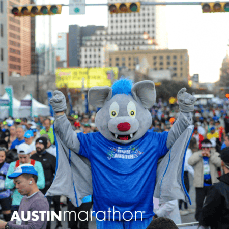 A large, costumed character dressed as a gray bat with a blue shirt that reads "Run Austin" raises its arms excitedly among a crowd of marathon runners in a city street. The text "AUSTIN marathon" is visible at the bottom of the image, capturing the vibrant spirit of marathon running. Austin Marathon Half Marathon & 5K A large, costumed character dressed as a gray bat with a blue shirt that reads "Run Austin" raises its arms excitedly among a crowd of marathon runners in a city street. The text "AUSTIN marathon" is visible at the bottom of the image, capturing the vibrant spirit of marathon running. Austin Marathon Half Marathon & 5K
