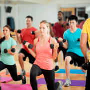 A group of people in a fitness class performing lunges with dumbbells, all aiming to achieve their marathon goals. They are on colorful exercise mats in a gym setting, dressed in athletic wear, and focusing on their workout in front of a large mirror. Austin Marathon Half Marathon & 5K
