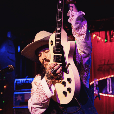 A guitarist dressed in a hat and patterned shirt plays passionately on stage, raising his white electric guitar above his head. Behind him, the red and neon-lit backdrop creates an energetic and lively atmosphere reminiscent of South Congress nights filled with vibrant eats and eclectic shops. Austin Marathon Half Marathon & 5K