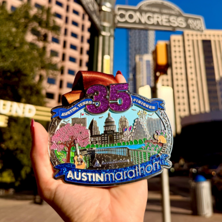 A hand holds a colorful 35th Austin Marathon belt buckle medal featuring unique medal designs and city landmarks, in front of downtown Austin buildings and a Congress Avenue street sign. Austin Marathon Half Marathon & 5K A hand holds a colorful 35th Austin Marathon belt buckle medal featuring unique medal designs and city landmarks, in front of downtown Austin buildings and a Congress Avenue street sign. Austin Marathon Half Marathon & 5K