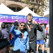 Two men, smiling and holding drinks, proudly display their medals. They stand in front of a tent with "Elite Light Lager" signage, amidst a buzzing crowd. It's race day at the Austin Marathon, and the lively scene suggests a celebratory outdoor event filled with marathon swag. Austin Marathon Half Marathon & 5K