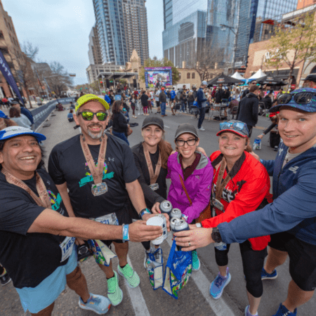 A group of six people in athletic gear celebrate with canned drinks on a city street during the 2025 Austin Marathon. They wear medals around their necks, surrounded by tall buildings and other jubilant race day participants. The mood is joyful and festive. Austin Marathon Half Marathon & 5K A group of six people in athletic gear celebrate with canned drinks on a city street during the 2025 Austin Marathon. They wear medals around their necks, surrounded by tall buildings and other jubilant race day participants. The mood is joyful and festive. Austin Marathon Half Marathon & 5K