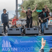 Amidst the vibrant excitement of Race Day, a band performs energetically on an outdoor stage. A female drummer and a singer with arms raised exude pure swag, while the guitarist and keyboard player groove nearby. A large banner in front proudly announces "Austin Marathon," as audience members enthusiastically raise their hands. Austin Marathon Half Marathon & 5K