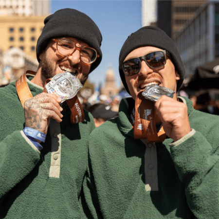 Two smiling runners in green fleeces and black beanies proudly bite medals around their necks, celebrating at a marathon in Austin with buildings and a lively crowd in the background. Austin Marathon Half Marathon & 5K Two smiling runners in green fleeces and black beanies proudly bite medals around their necks, celebrating at a marathon in Austin with buildings and a lively crowd in the background. Austin Marathon Half Marathon & 5K