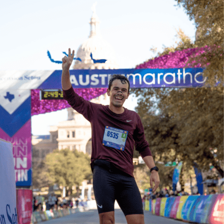 A smiling runner raises a peace sign as he crosses the finish line at the best marathon in Austin, with race bib 6535. The event banner and a dome-shaped building are visible in the background. Austin Marathon Half Marathon & 5K A smiling runner raises a peace sign as he crosses the finish line at the best marathon in Austin, with race bib 6535. The event banner and a dome-shaped building are visible in the background. Austin Marathon Half Marathon & 5K