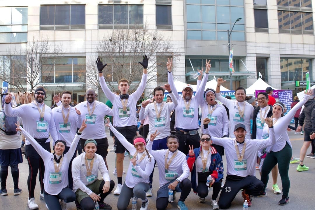 A group of marathon runners from corporate teams, wearing matching white shirts, pose together, smiling and raising their arms in celebration with medals around their necks. A city building and other participants are visible in the background. Austin Marathon Half Marathon & 5K