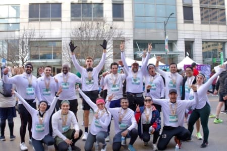 A group of marathon runners from corporate teams, wearing matching white shirts, pose together, smiling and raising their arms in celebration with medals around their necks. A city building and other participants are visible in the background. Austin Marathon Half Marathon & 5K