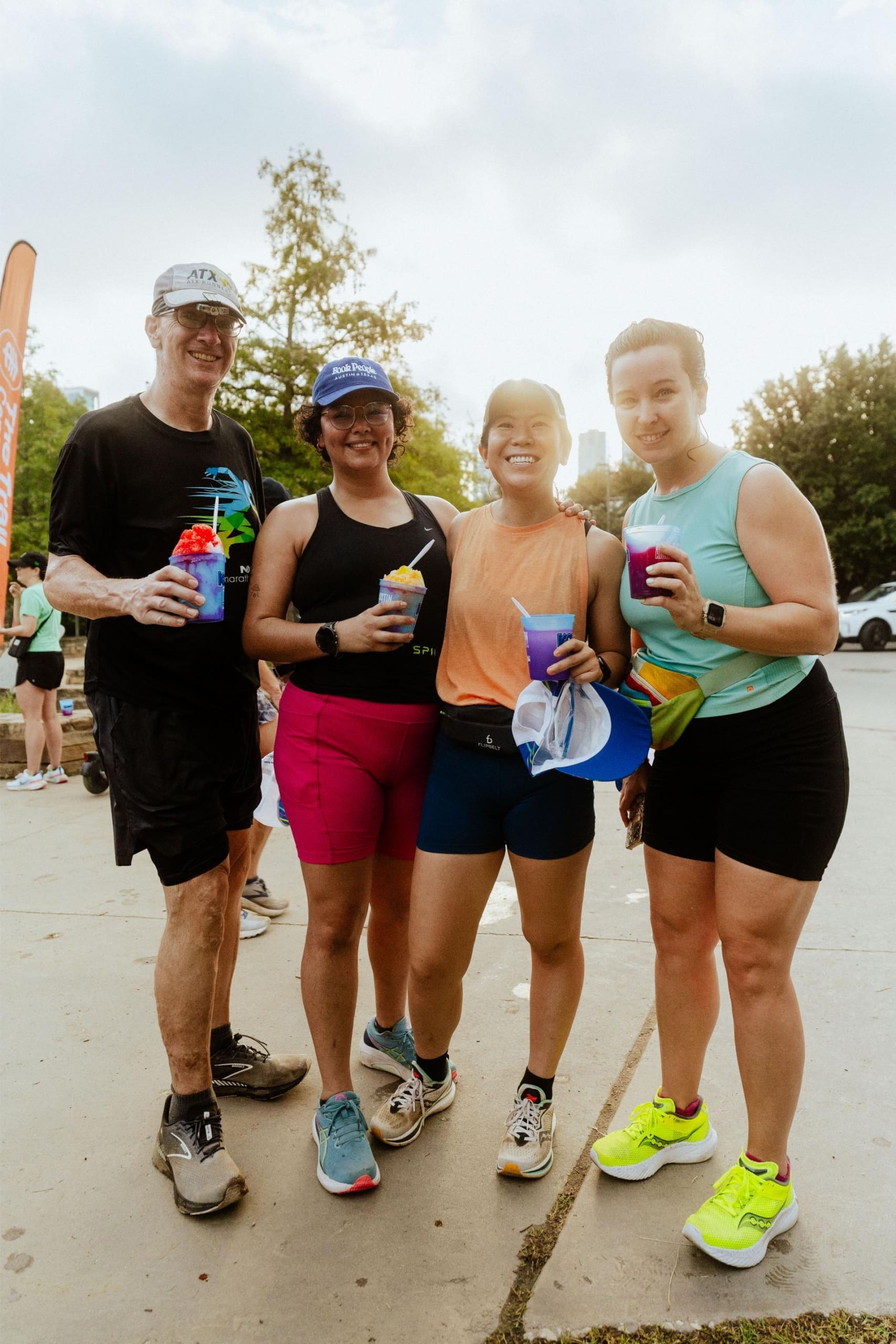 Four people in athletic wear stand outdoors, smiling and holding colorful drinks—Snow Cones on Trail—suggesting they just finished a run or race. Trees and fellow participants can be seen in the background. Austin Marathon Half Marathon & 5K Four people in athletic wear stand outdoors, smiling and holding colorful drinks—Snow Cones on Trail—suggesting they just finished a run or race. Trees and fellow participants can be seen in the background. Austin Marathon Half Marathon & 5K