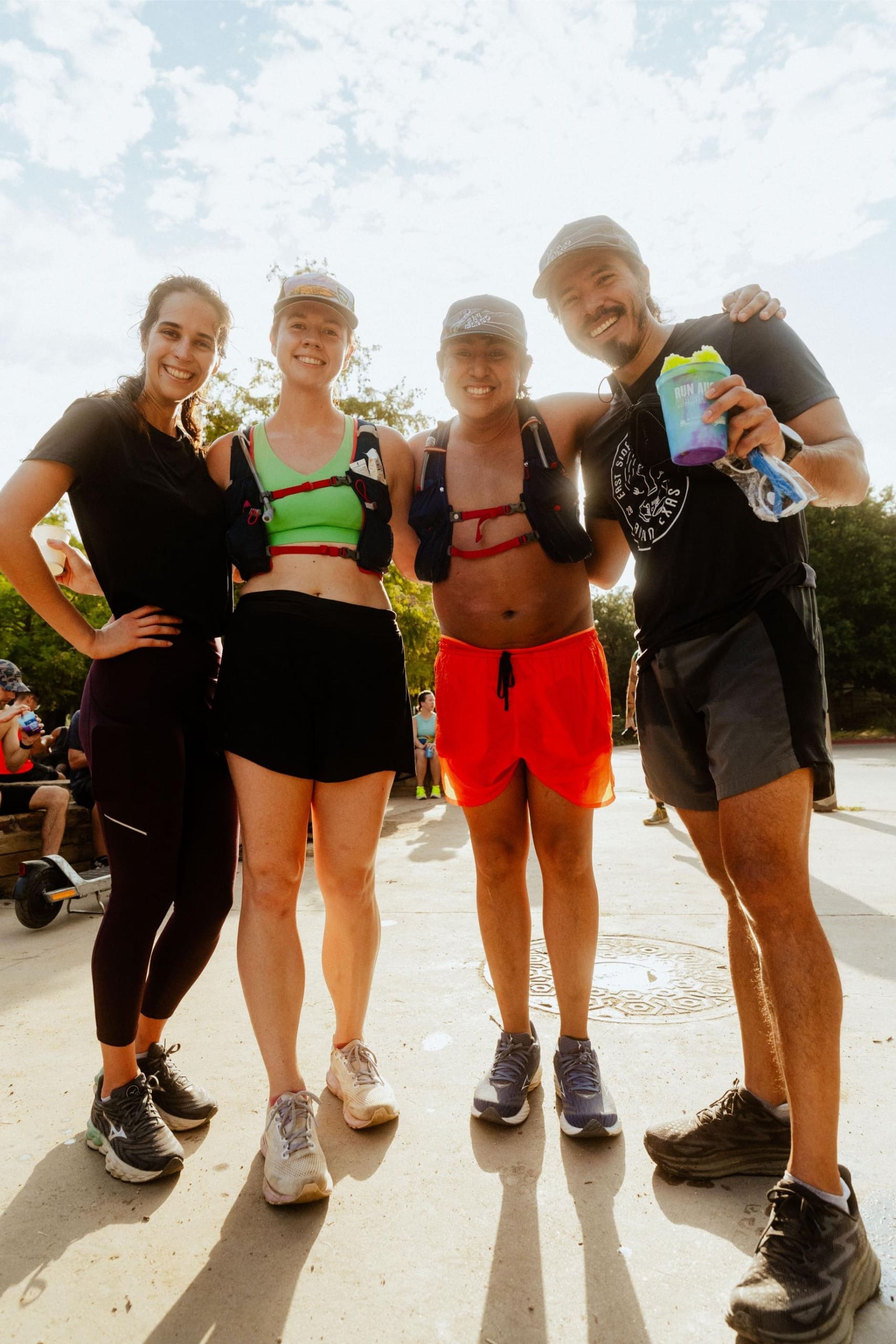 Four smiling people in athletic wear stand close together outdoors on a sunny day, appearing happy after a run or workout—perhaps celebrating with snow cones on trail. Trees and other people are visible in the background. Austin Marathon Half Marathon & 5K Four smiling people in athletic wear stand close together outdoors on a sunny day, appearing happy after a run or workout—perhaps celebrating with snow cones on trail. Trees and other people are visible in the background. Austin Marathon Half Marathon & 5K
