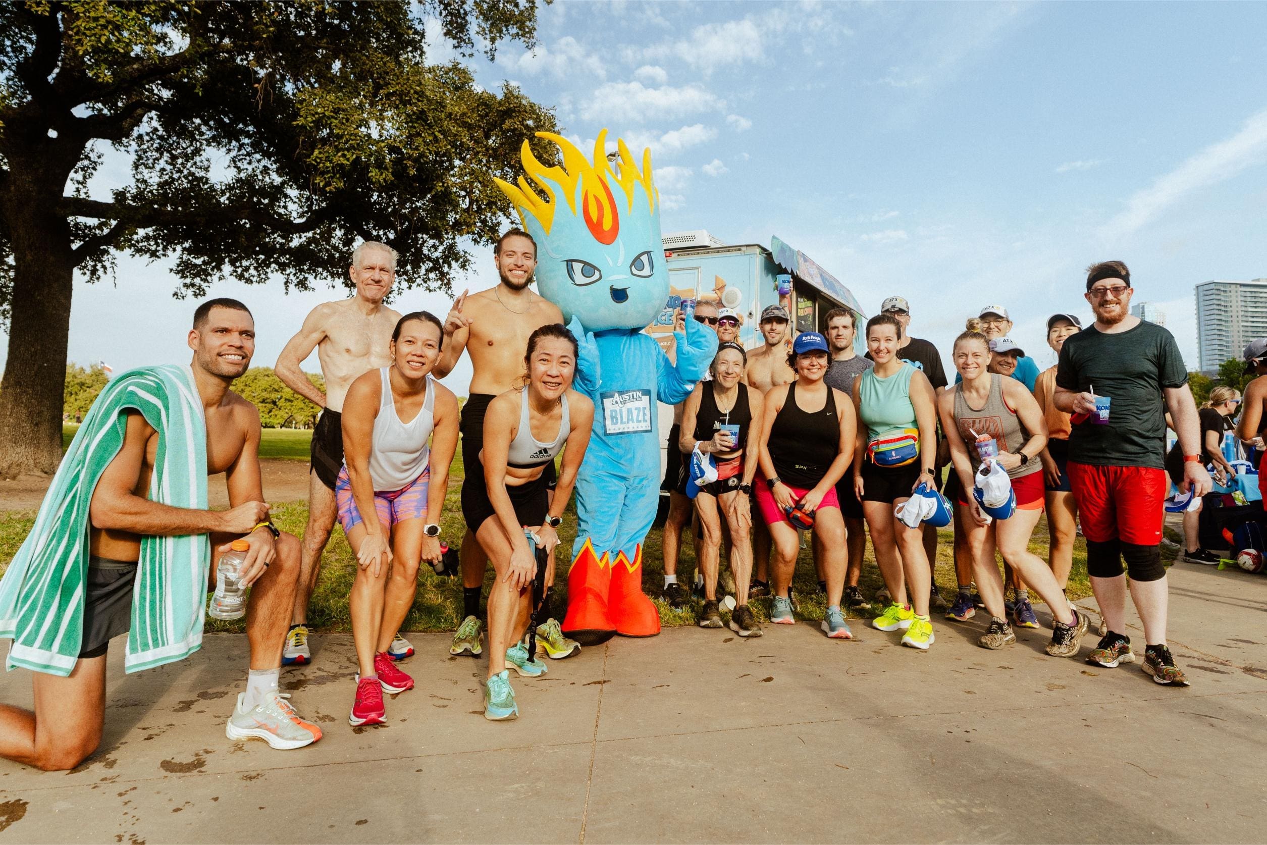 A group of smiling runners poses outdoors with a person in a blue flame mascot costume. Clutching water bottles and enjoying Snow Cones on Trail, they stand in athletic gear with trees and buildings in the background on a sunny day. Austin Marathon Half Marathon & 5K A group of smiling runners poses outdoors with a person in a blue flame mascot costume. Clutching water bottles and enjoying Snow Cones on Trail, they stand in athletic gear with trees and buildings in the background on a sunny day. Austin Marathon Half Marathon & 5K