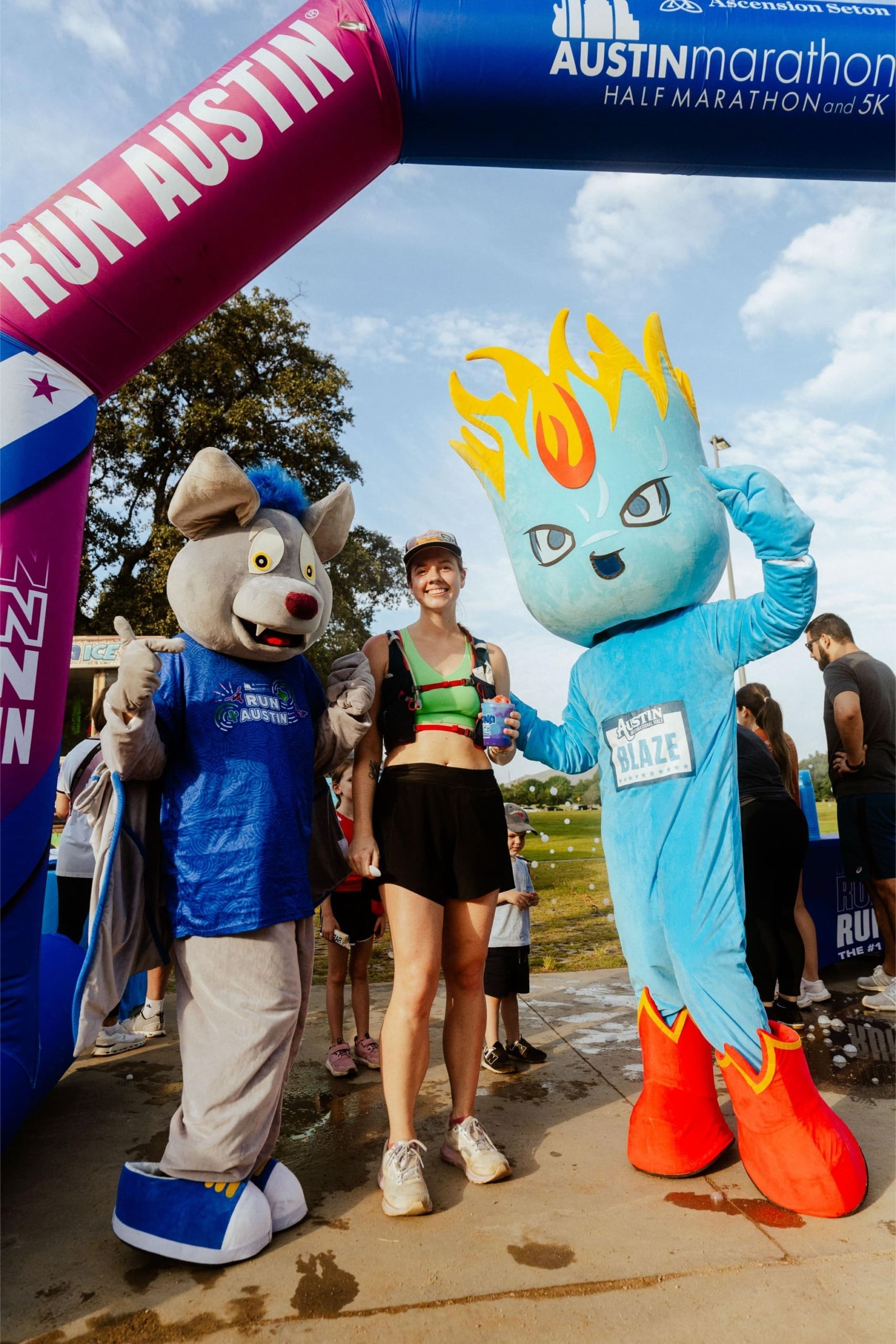 A smiling runner stands between two colorful mascots at the finish line of the Austin Marathon, under an arch that reads "RUN AUSTIN," with Snow Cones on Trail nearby. Other people and trees are visible in the background. Austin Marathon Half Marathon & 5K A smiling runner stands between two colorful mascots at the finish line of the Austin Marathon, under an arch that reads "RUN AUSTIN," with Snow Cones on Trail nearby. Other people and trees are visible in the background. Austin Marathon Half Marathon & 5K