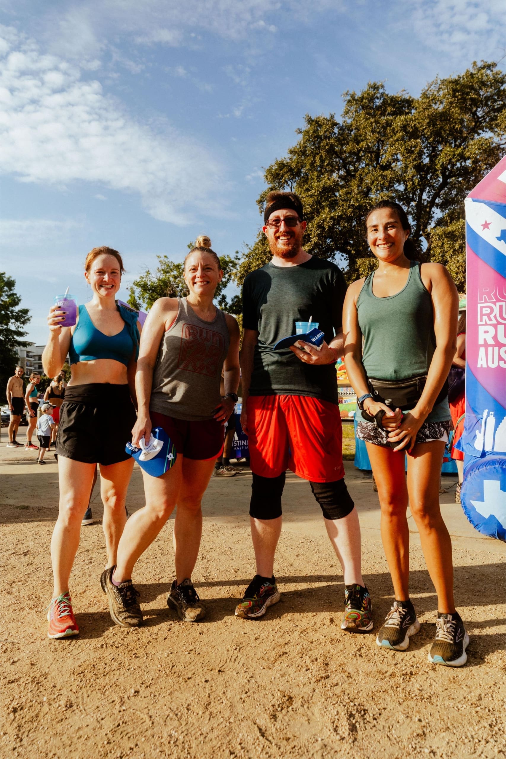 Four runners stand smiling together outdoors at a race event, holding water bottles. They wear athletic clothes and sneakers, with trees, a blue sky, and Snow Cones on Trail banners visible among other participants in the background. Austin Marathon Half Marathon & 5K Four runners stand smiling together outdoors at a race event, holding water bottles. They wear athletic clothes and sneakers, with trees, a blue sky, and Snow Cones on Trail banners visible among other participants in the background. Austin Marathon Half Marathon & 5K