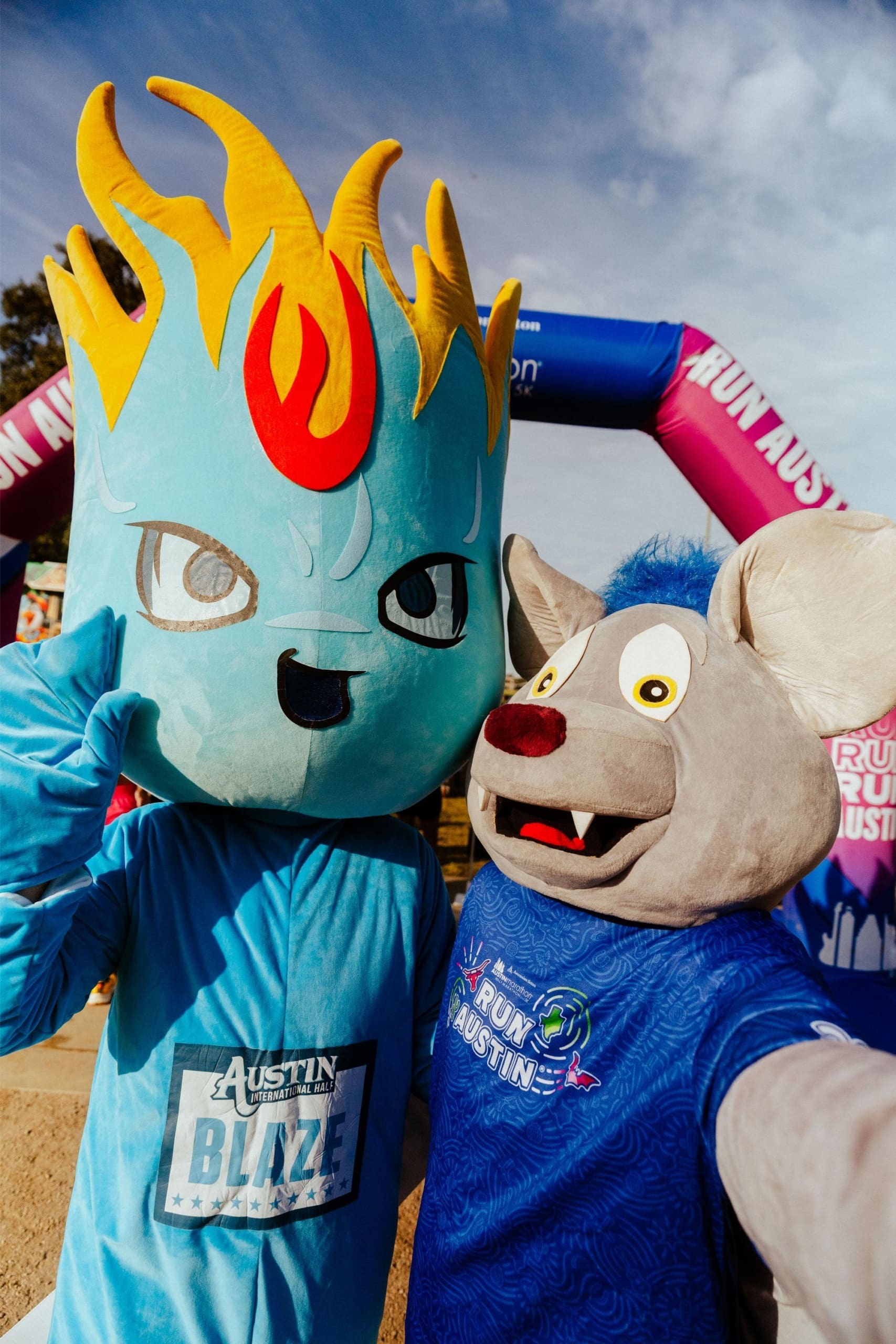 Two mascots in costume pose for a selfie at an outdoor event, with vibrant arches behind them. The blue flame-haired mascot and the gray mouse in a blue shirt celebrate the fun of Snow Cones on Trail. Austin Marathon Half Marathon & 5K Two mascots in costume pose for a selfie at an outdoor event, with vibrant arches behind them. The blue flame-haired mascot and the gray mouse in a blue shirt celebrate the fun of Snow Cones on Trail. Austin Marathon Half Marathon & 5K