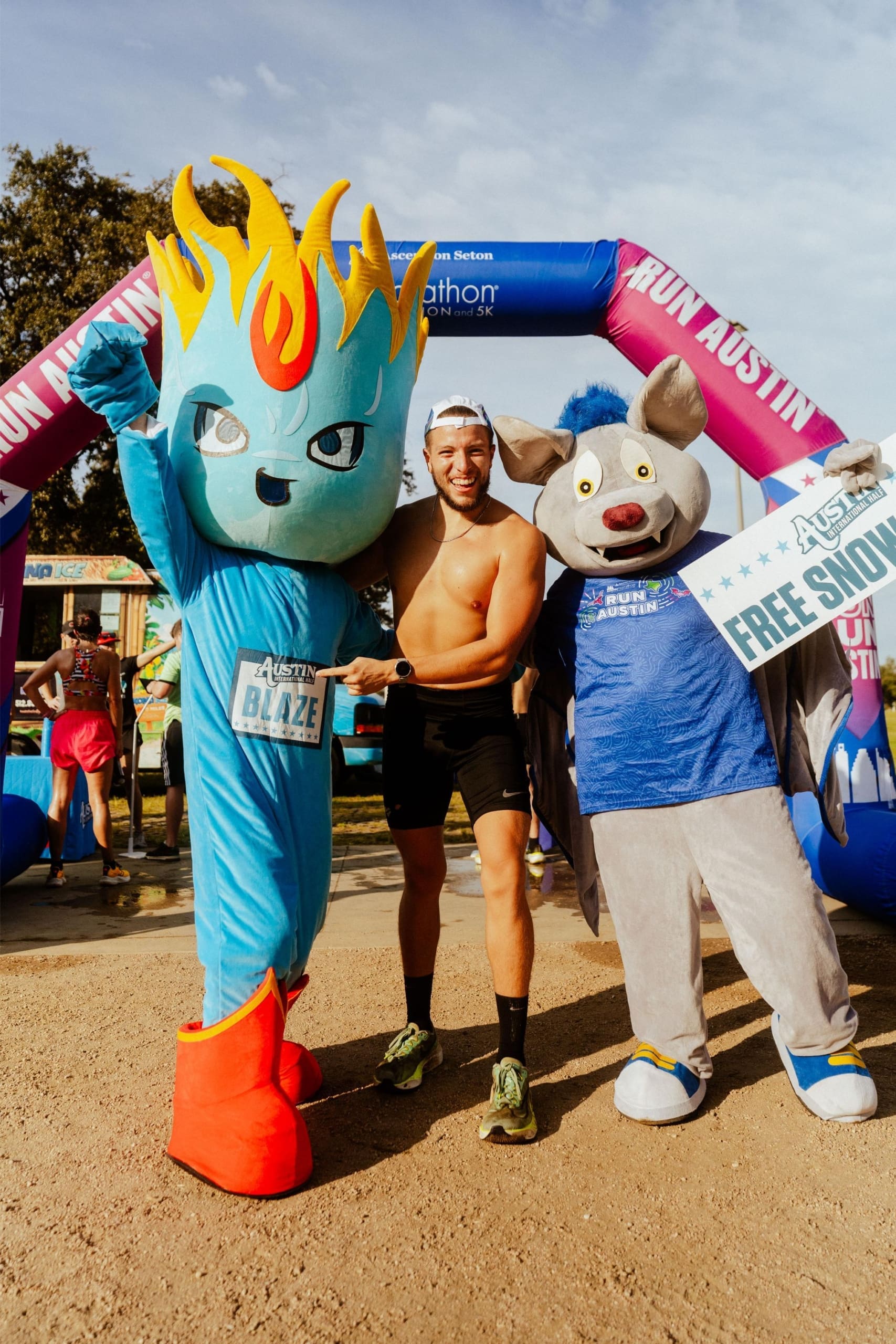 A smiling runner poses between two mascots in costume at a race finish line, celebrating with Snow Cones on Trail. One mascot is blue with a flame head, the other is a gray bat holding a "FREE SQUAD" sign. An inflatable arch and trees are in the background. Austin Marathon Half Marathon & 5K A smiling runner poses between two mascots in costume at a race finish line, celebrating with Snow Cones on Trail. One mascot is blue with a flame head, the other is a gray bat holding a "FREE SQUAD" sign. An inflatable arch and trees are in the background. Austin Marathon Half Marathon & 5K