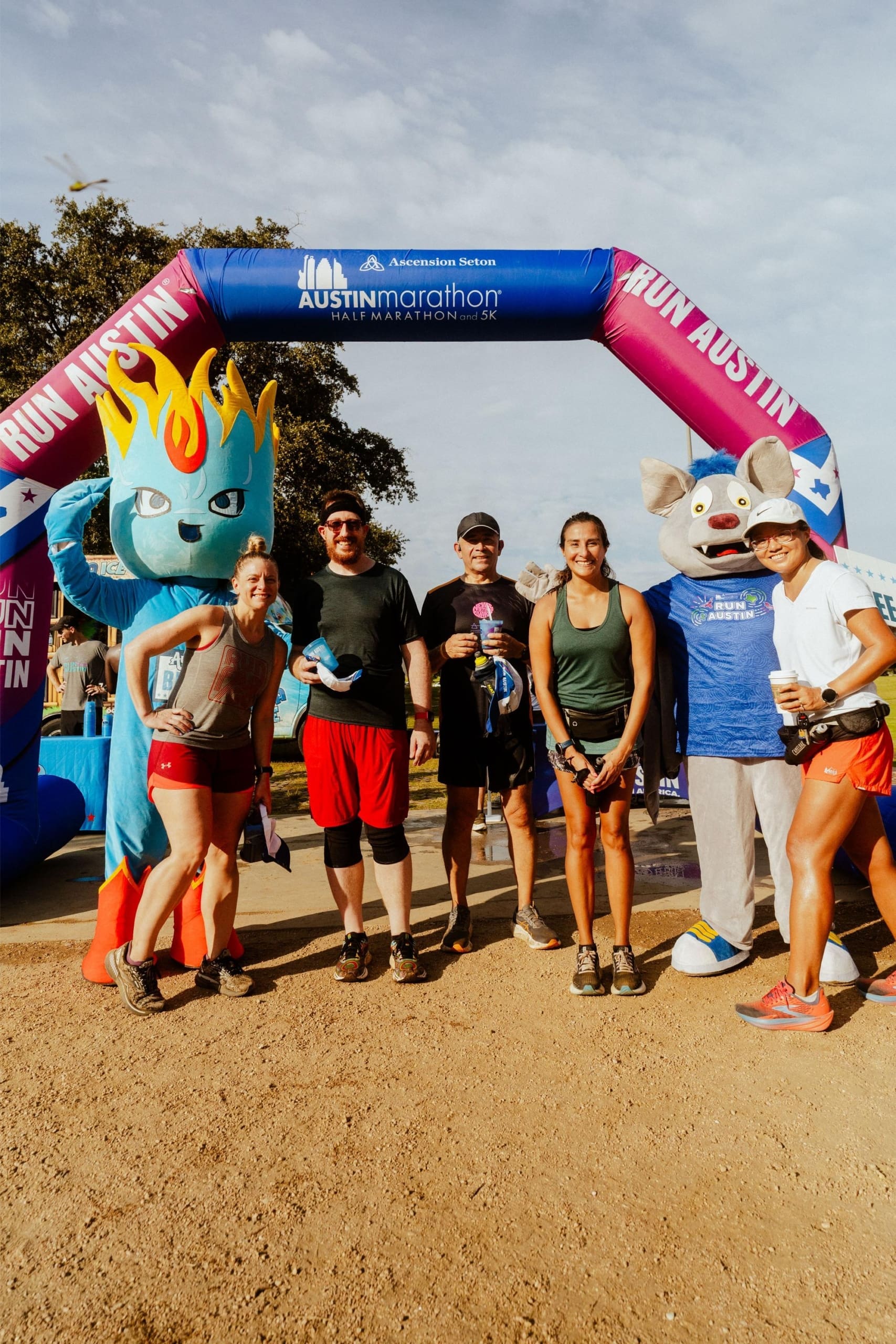 A group of marathon runners pose smiling under an Austin Marathon archway, joined by two mascots—one a blue flame character, the other a gray animal in a blue shirt—with Snow Cones on Trail stands nearby. The sun is shining and trees fill the background. Austin Marathon Half Marathon & 5K A group of marathon runners pose smiling under an Austin Marathon archway, joined by two mascots—one a blue flame character, the other a gray animal in a blue shirt—with Snow Cones on Trail stands nearby. The sun is shining and trees fill the background. Austin Marathon Half Marathon & 5K