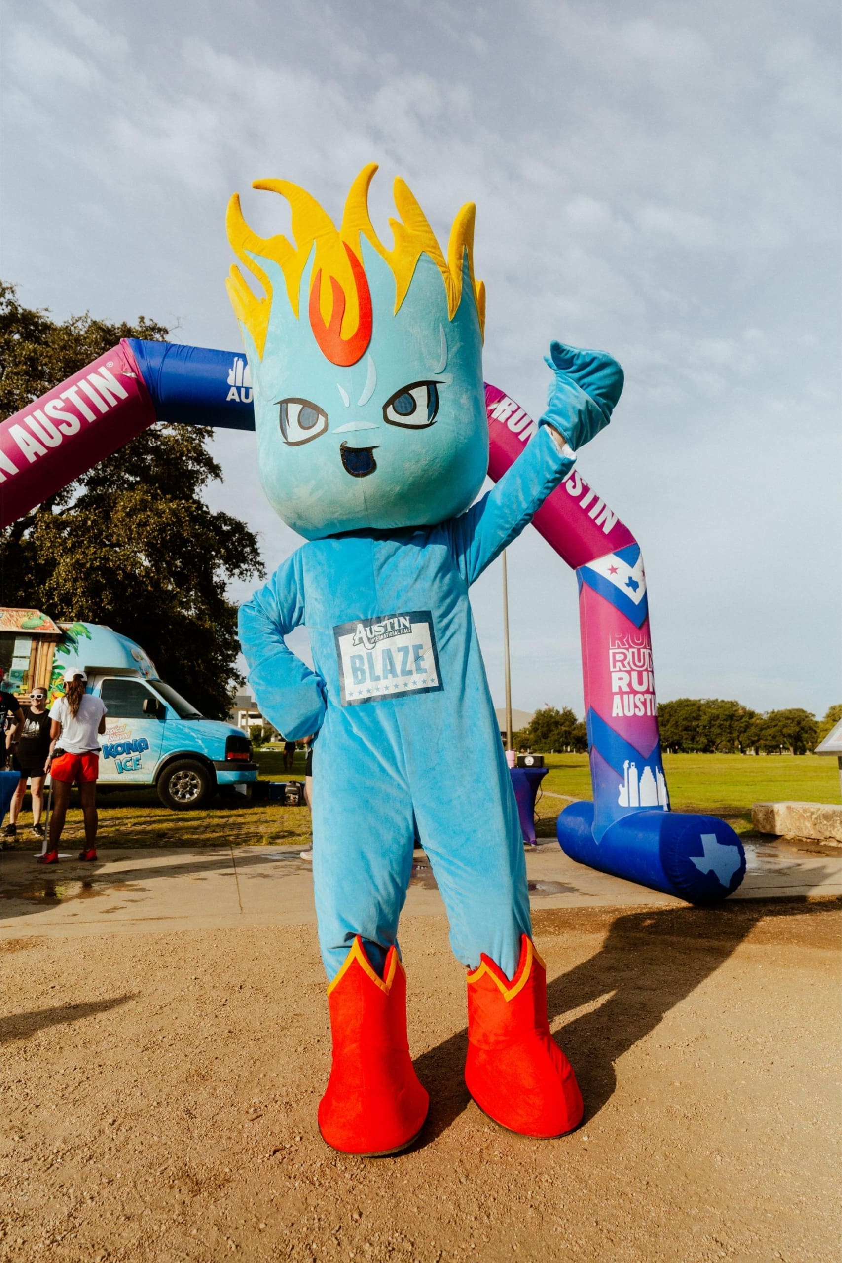 A person in a blue flame-themed mascot costume labeled "Austin Blaze" poses with one arm raised at an outdoor event, standing by a colorful inflatable arch and the Snow Cones on Trail ice cream truck. Austin Marathon Half Marathon & 5K A person in a blue flame-themed mascot costume labeled "Austin Blaze" poses with one arm raised at an outdoor event, standing by a colorful inflatable arch and the Snow Cones on Trail ice cream truck. Austin Marathon Half Marathon & 5K