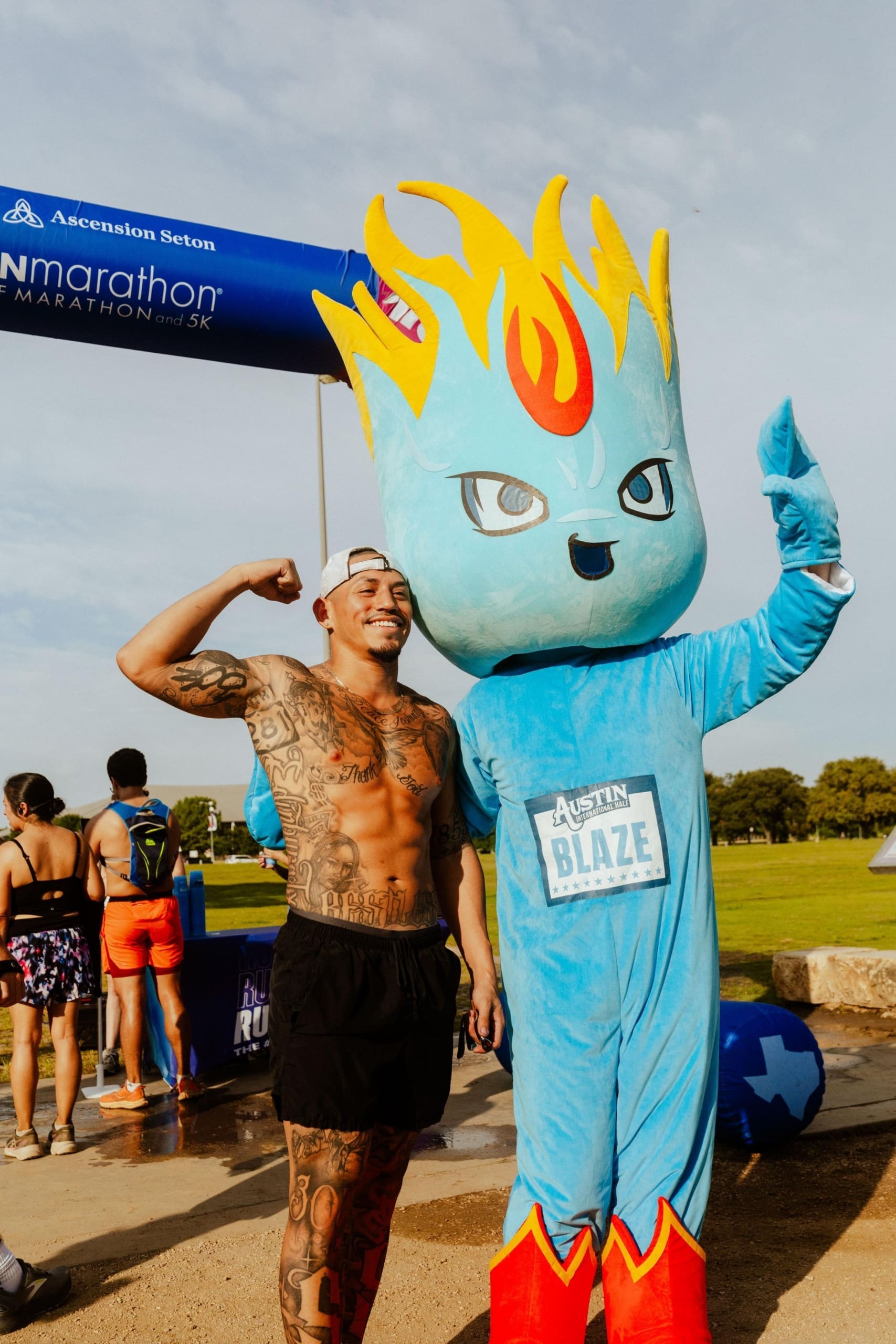 A shirtless, tattooed man flexes his arm and smiles beside the Austin Blaze mascot—blue-bodied with fiery yellow hair—at a marathon event finish line, where participants celebrate and enjoy Snow Cones on Trail in the background. Austin Marathon Half Marathon & 5K A shirtless, tattooed man flexes his arm and smiles beside the Austin Blaze mascot—blue-bodied with fiery yellow hair—at a marathon event finish line, where participants celebrate and enjoy Snow Cones on Trail in the background. Austin Marathon Half Marathon & 5K