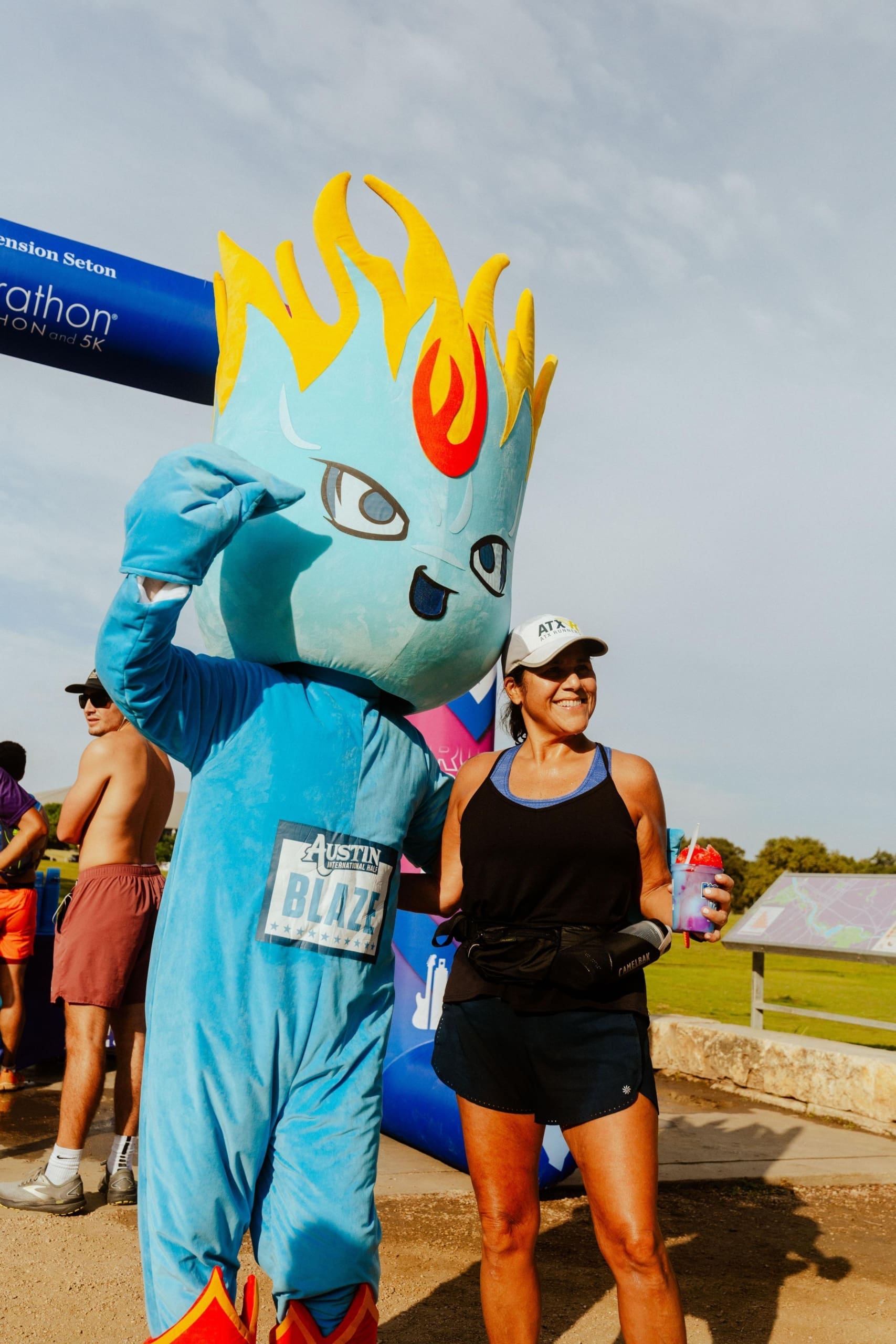 A person in a flame-headed blue mascot costume poses with a smiling woman in athletic gear near the marathon finish line, where runners enjoyed Snow Cones on Trail at the outdoor event. Austin Marathon Half Marathon & 5K A person in a flame-headed blue mascot costume poses with a smiling woman in athletic gear near the marathon finish line, where runners enjoyed Snow Cones on Trail at the outdoor event. Austin Marathon Half Marathon & 5K