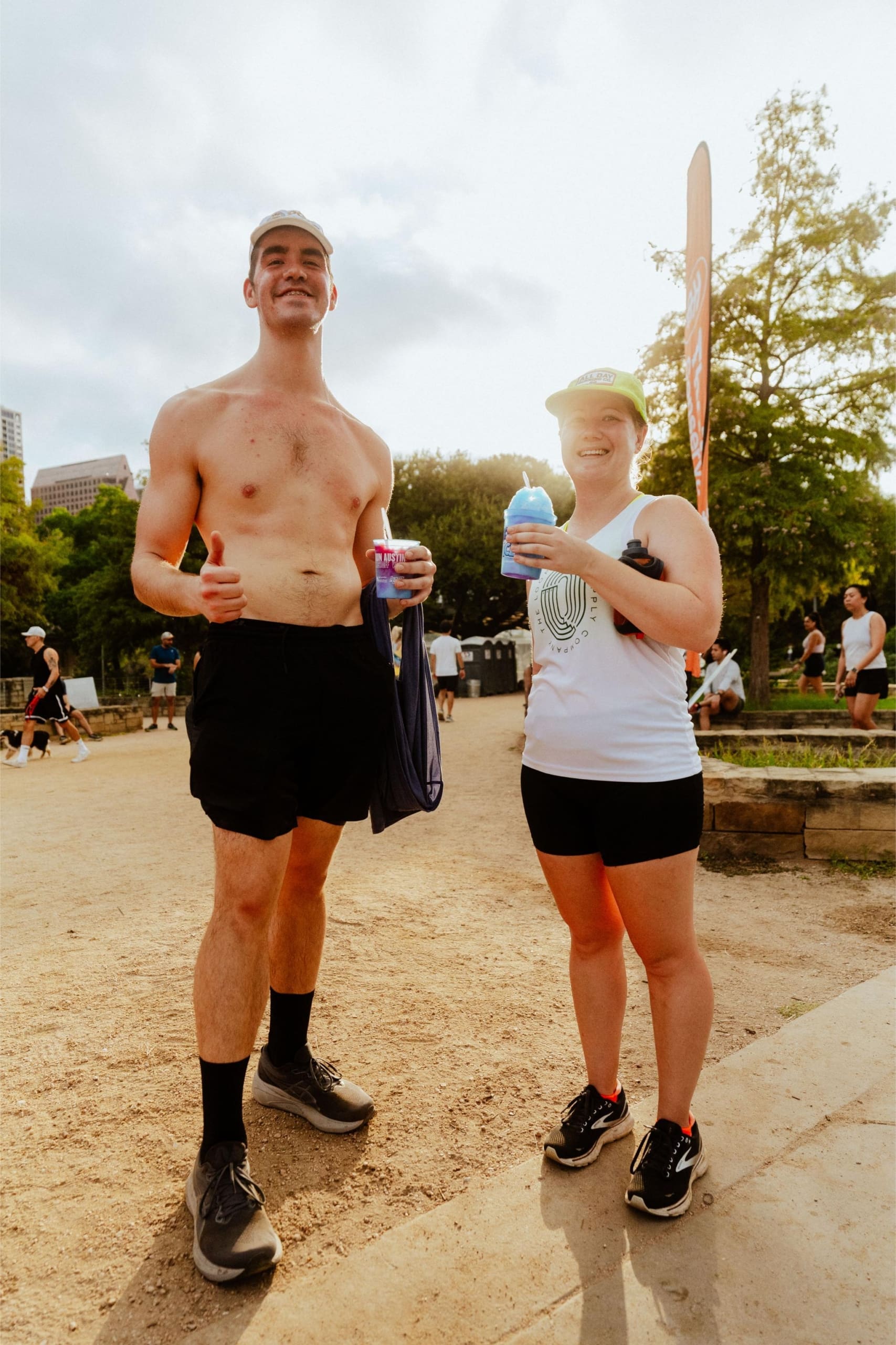 A man and woman in athletic wear stand outdoors on a dirt path, smiling and holding snow cones on trail. The man is shirtless, and both wear hats. Other people and trees are visible in the background on a sunny day. Austin Marathon Half Marathon & 5K A man and woman in athletic wear stand outdoors on a dirt path, smiling and holding snow cones on trail. The man is shirtless, and both wear hats. Other people and trees are visible in the background on a sunny day. Austin Marathon Half Marathon & 5K