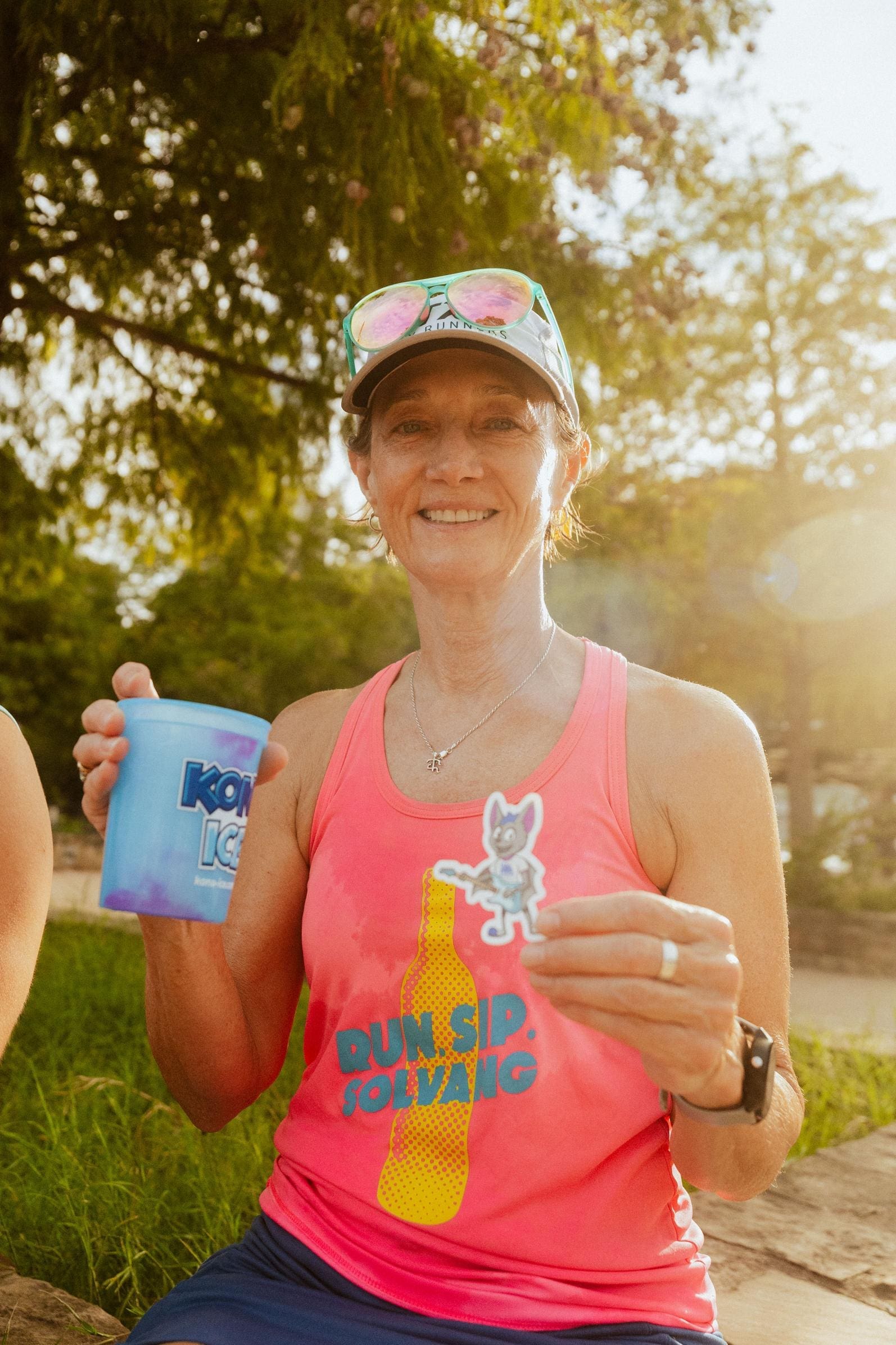 A smiling woman in a pink tank top and cap holds a blue cup and a small cartoon cat sticker, enjoying Snow Cones on Trail outdoors on a sunny day. She wears sunglasses on her head and sits on grass with trees behind her. Austin Marathon Half Marathon & 5K A smiling woman in a pink tank top and cap holds a blue cup and a small cartoon cat sticker, enjoying Snow Cones on Trail outdoors on a sunny day. She wears sunglasses on her head and sits on grass with trees behind her. Austin Marathon Half Marathon & 5K