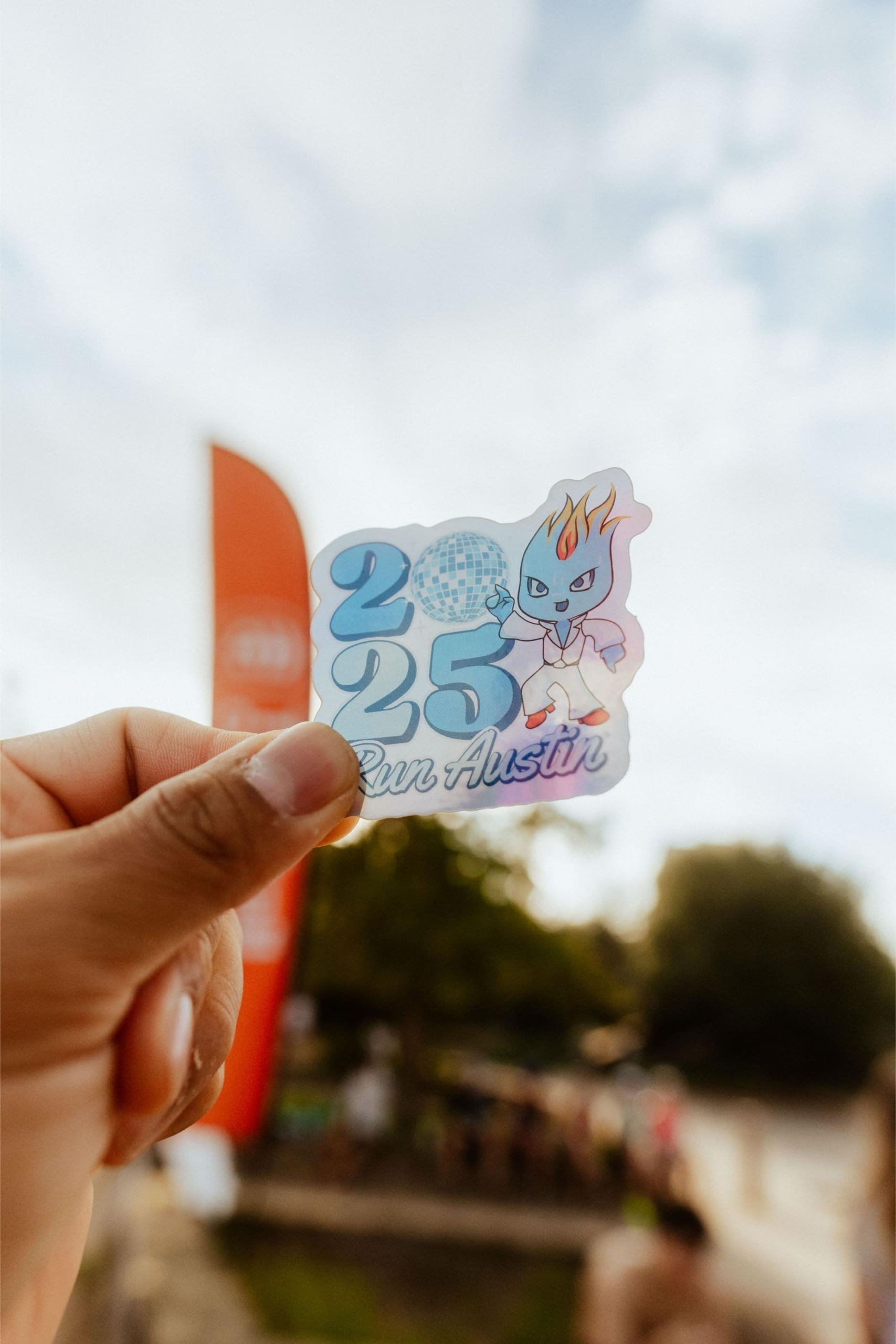 A hand holds a sticker that reads “2025 Run Austin” with a cartoon character and disco ball graphic, against a blurred outdoor background with trees, a red vertical banner, and hints of Snow Cones on Trail festivities. Austin Marathon Half Marathon & 5K A hand holds a sticker that reads “2025 Run Austin” with a cartoon character and disco ball graphic, against a blurred outdoor background with trees, a red vertical banner, and hints of Snow Cones on Trail festivities. Austin Marathon Half Marathon & 5K
