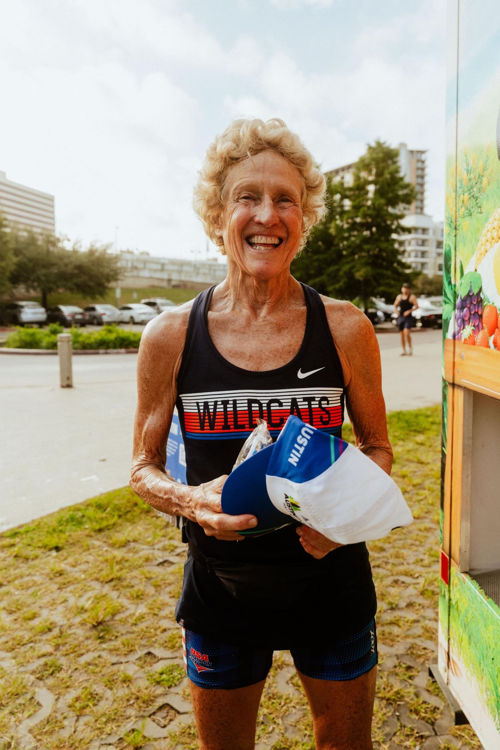 An older woman with curly blonde hair, wearing a "WILDCATS" running tank, smiles while holding a blue and white cap outdoors on a sunny day, enjoying Snow Cones on Trail with buildings and greenery in the background. Austin Marathon Half Marathon & 5K An older woman with curly blonde hair, wearing a "WILDCATS" running tank, smiles while holding a blue and white cap outdoors on a sunny day, enjoying Snow Cones on Trail with buildings and greenery in the background. Austin Marathon Half Marathon & 5K