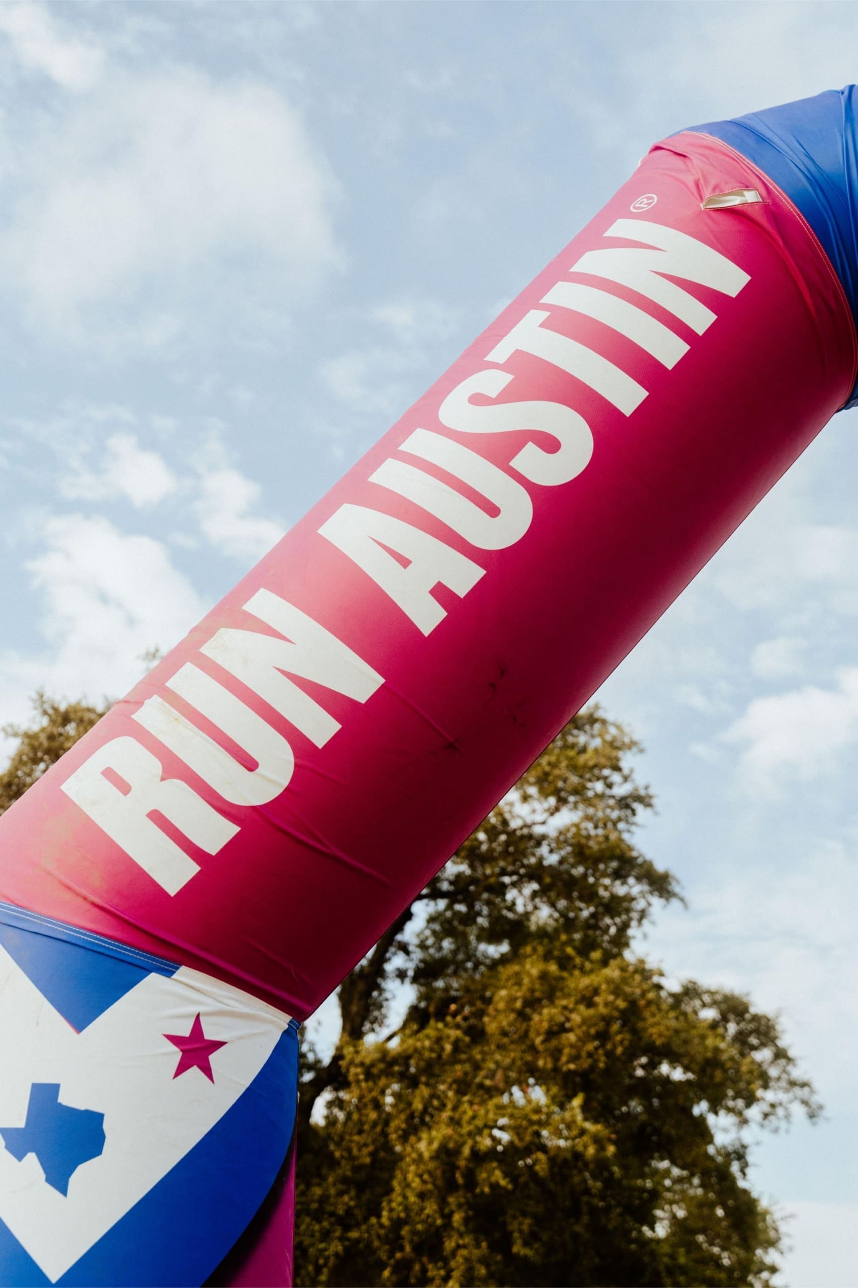 A large pink and blue inflatable arch with the words "RUN AUSTIN" in bold white letters stands outdoors beneath a blue sky, inviting runners to enjoy Snow Cones on Trail amid the trees. Austin Marathon Half Marathon & 5K A large pink and blue inflatable arch with the words "RUN AUSTIN" in bold white letters stands outdoors beneath a blue sky, inviting runners to enjoy Snow Cones on Trail amid the trees. Austin Marathon Half Marathon & 5K
