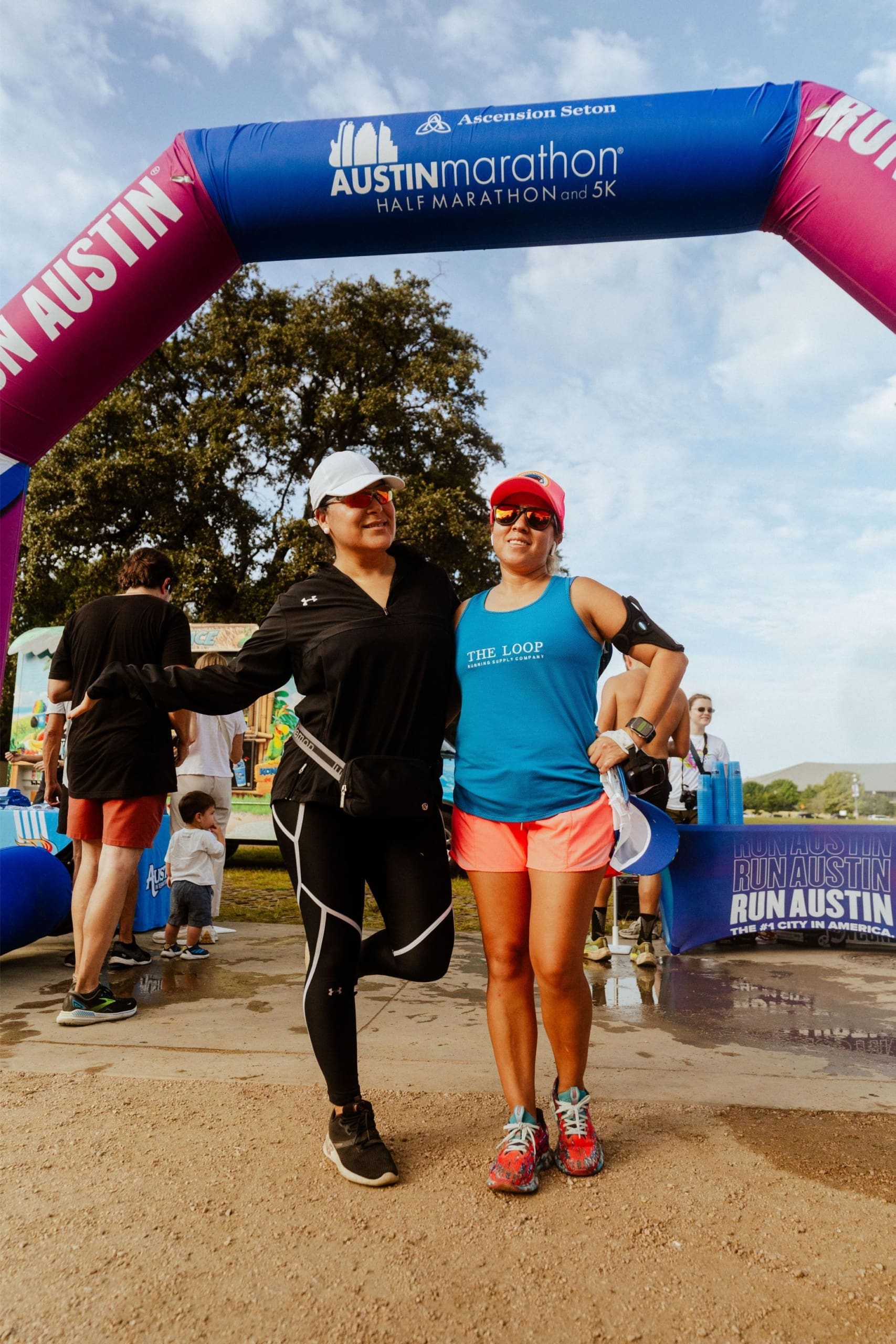 Two women in athletic wear smile and pose together under a purple and blue “Austin Marathon” archway at the finish line, with other participants, a child, and Snow Cones on Trail stands visible in the background. Austin Marathon Half Marathon & 5K Two women in athletic wear smile and pose together under a purple and blue “Austin Marathon” archway at the finish line, with other participants, a child, and Snow Cones on Trail stands visible in the background. Austin Marathon Half Marathon & 5K