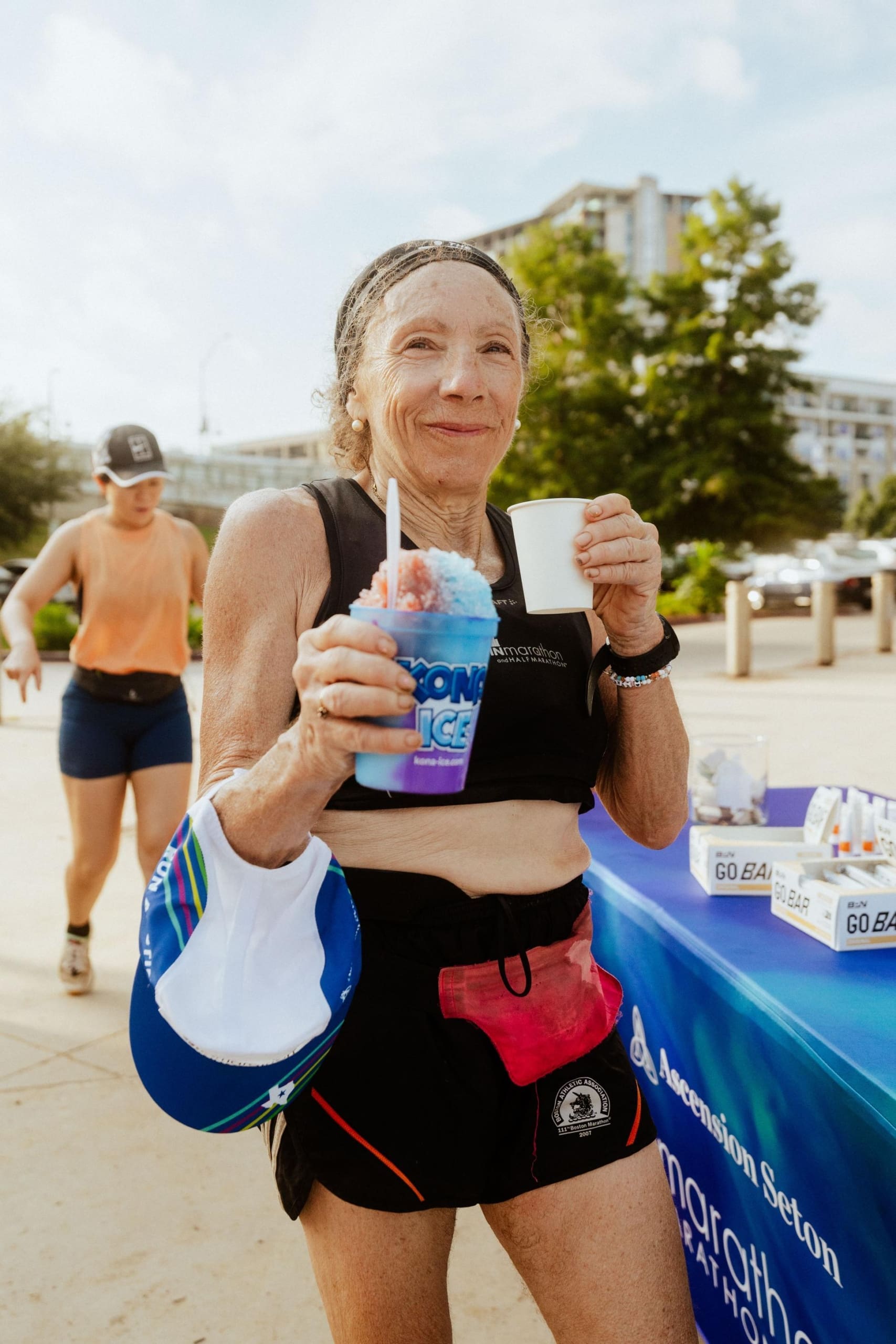 An older woman in athletic gear smiles while holding a snow cone and a cup, standing outdoors near a table with snacks and drinks. Another person in athletic wear walks behind her—enjoying Snow Cones on Trail on a sunny day. Austin Marathon Half Marathon & 5K An older woman in athletic gear smiles while holding a snow cone and a cup, standing outdoors near a table with snacks and drinks. Another person in athletic wear walks behind her—enjoying Snow Cones on Trail on a sunny day. Austin Marathon Half Marathon & 5K