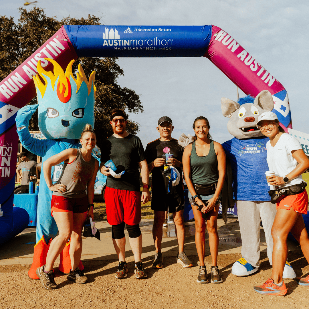 Six smiling runners pose under a colorful "Run Austin" arch, flanked by two mascots—one blue with a flame head and another gray with large ears. Everyone looks happy, dressed in athletic gear, ready to enjoy Snow Cones on Trail after the race. Austin Marathon Half Marathon & 5K Six smiling runners pose under a colorful "Run Austin" arch, flanked by two mascots—one blue with a flame head and another gray with large ears. Everyone looks happy, dressed in athletic gear, ready to enjoy Snow Cones on Trail after the race. Austin Marathon Half Marathon & 5K