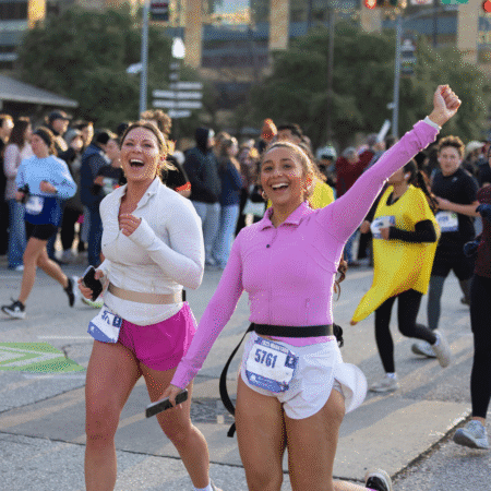 Two women in athletic clothes run and smile at a race, both with bib numbers and one raising her arm in excitement. The crowd of runners behind them, including someone in a banana costume, highlights the lively spirit of this training base event. Austin Marathon Half Marathon & 5K