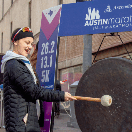 A woman in athletic clothing smiles while striking a large gong with a mallet at the Austin Marathon event, providing more reasons to get excited for 2026. A race sign showing distances is visible in the background. Austin Marathon Half Marathon & 5K A woman in athletic clothing smiles while striking a large gong with a mallet at the Austin Marathon event, providing more reasons to get excited for 2026. A race sign showing distances is visible in the background. Austin Marathon Half Marathon & 5K