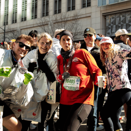 A group of marathon runners smiling and posing with their medals and drinks after finishing the 2026 Austin Marathon, with others cheering in the background on a city street. Austin Marathon Half Marathon & 5K