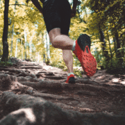 A person wearing black shorts and red running shoes is trail running through a sunlit forest in Austin, stepping over exposed tree roots on a dirt path. The focus is on their legs and shoes—a perfect scene for a beginner's guide to trail running. Austin Marathon Half Marathon & 5K