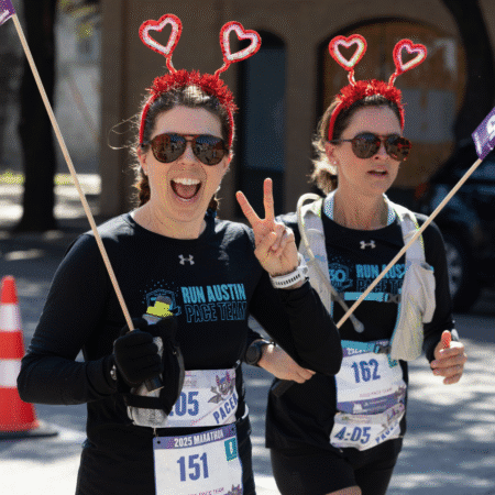 Two women running the Austin Marathon, wearing black "Run Austin Pace Team" shirts and fun red heart-shaped headbands. One smiles and flashes a peace sign; both hold small flags and have race bibs pinned to their outfits. Austin Marathon Half Marathon & 5K