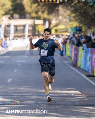 A male runner in a navy shirt and shorts crosses the finish line at the Austin Marathon race, arms raised in celebration, with confetti on the ground and spectators lining colorful barriers, ready to capture personal official race photos. Austin Marathon Half Marathon & 5K A male runner in a navy shirt and shorts crosses the finish line at the Austin Marathon race, arms raised in celebration, with confetti on the ground and spectators lining colorful barriers, ready to capture personal official race photos. Austin Marathon Half Marathon & 5K