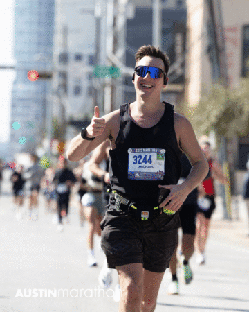 A smiling runner wearing sunglasses, a bib labeled "3244 Michael," and a black tank top gives a thumbs-up during the 2023 Austin Marathon race. Other runners and city buildings appear in the background, creating a lively scene perfect for Official Race Photos. Austin Marathon Half Marathon & 5K A smiling runner wearing sunglasses, a bib labeled "3244 Michael," and a black tank top gives a thumbs-up during the 2023 Austin Marathon race. Other runners and city buildings appear in the background, creating a lively scene perfect for Official Race Photos. Austin Marathon Half Marathon & 5K