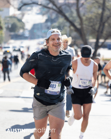 A smiling man in a dark shirt and cap runs in the Austin Marathon race, wearing bib number 1483. Other runners and a sunny, tree-lined street are visible in the background. This moment captures an inspiring personal achievement. Austin Marathon Half Marathon & 5K A smiling man in a dark shirt and cap runs in the Austin Marathon race, wearing bib number 1483. Other runners and a sunny, tree-lined street are visible in the background. This moment captures an inspiring personal achievement. Austin Marathon Half Marathon & 5K