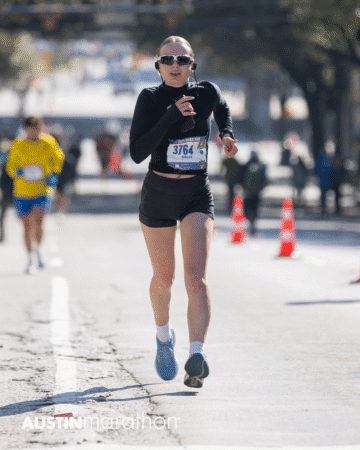 A runner wearing sunglasses, a black long-sleeve shirt, and shorts, with bib number 3764, competes in the Austin Marathon race. Other runners and orange traffic cones are visible in this Official Race Photo background. Austin Marathon Half Marathon & 5K A runner wearing sunglasses, a black long-sleeve shirt, and shorts, with bib number 3764, competes in the Austin Marathon race. Other runners and orange traffic cones are visible in this Official Race Photo background. Austin Marathon Half Marathon & 5K