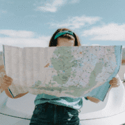 A person stands outdoors in front of a car, holding and looking at a large unfolded map that covers their face, with a blue sky and clouds in the background—like a beginner's guide to trail running adventure. Austin Marathon Half Marathon & 5K