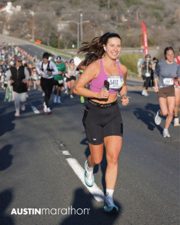 A woman wearing a race bib runs and smiles during the Austin Marathon, with a crowd of other runners behind her on a sunny road. The event logo "AUSTINmarathon" is visible in the bottom left corner, capturing an Official Race Photo. Austin Marathon Half Marathon & 5K A woman wearing a race bib runs and smiles during the Austin Marathon, with a crowd of other runners behind her on a sunny road. The event logo "AUSTINmarathon" is visible in the bottom left corner, capturing an Official Race Photo. Austin Marathon Half Marathon & 5K