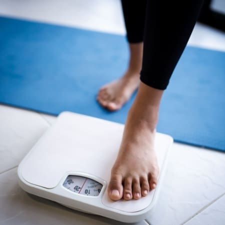 A person stands with one bare foot on a white bathroom scale, next to a blue exercise mat on a tiled floor, perhaps curious about the running calories or calories burned during their workout. The scale's dial is partially visible. Austin Marathon Half Marathon & 5K