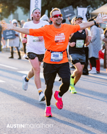 A smiling man in an orange shirt and pink shoes runs with arms outstretched at the Austin Marathon, capturing a personal moment during the race, surrounded by other runners and spectators in the background. Austin Marathon Half Marathon & 5K A smiling man in an orange shirt and pink shoes runs with arms outstretched at the Austin Marathon, capturing a personal moment during the race, surrounded by other runners and spectators in the background. Austin Marathon Half Marathon & 5K