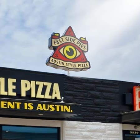 A rooftop sign for "East Side Pies: Austin Style Pizza," one of the best Austin restaurants, features a yellow eye in a red triangle above a black and white building facade. Part of a yellow "PIZZA" sign is visible below. Austin Marathon Half Marathon & 5K