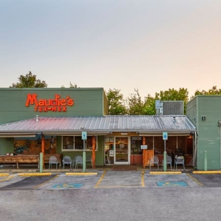 A green building with a metal roof houses Maudie’s Tex-Mex restaurant, a popular Austin spot for carb-loading. The front features a red neon sign, outdoor seating with tables and chairs, handicap parking spaces, and trees in the background. Austin Marathon Half Marathon & 5K