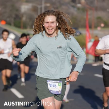 A smiling man with long curly hair, wearing a light green shirt and shorts, runs in a marathon with a race bib numbered 6755, burning calories as he goes. Other runners are visible in the background on this sunny day. Austin Marathon Half Marathon & 5K A smiling man with long curly hair, wearing a light green shirt and shorts, runs in a marathon with a race bib numbered 6755, burning calories as he goes. Other runners are visible in the background on this sunny day. Austin Marathon Half Marathon & 5K